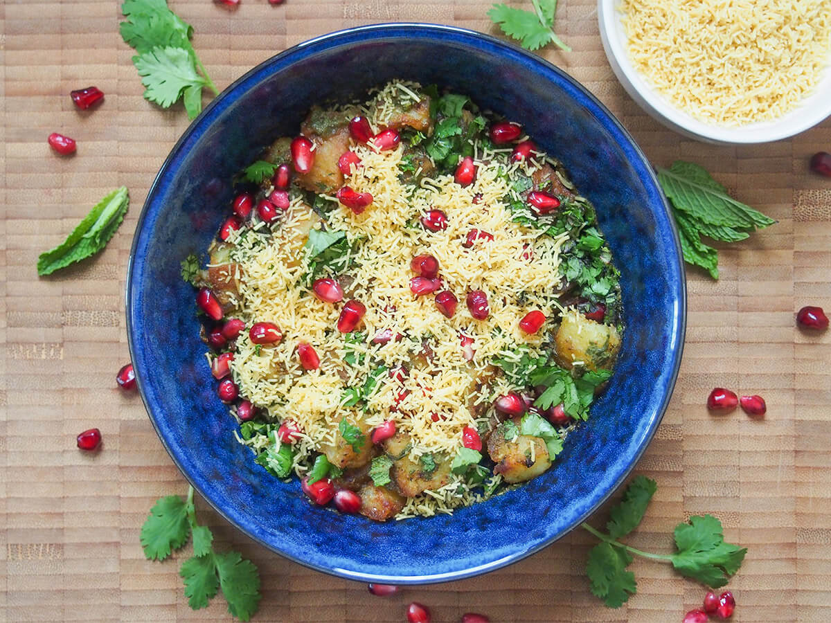 bowl of aloo chaat with additional sev to side and herbs and pomegranate around bowl