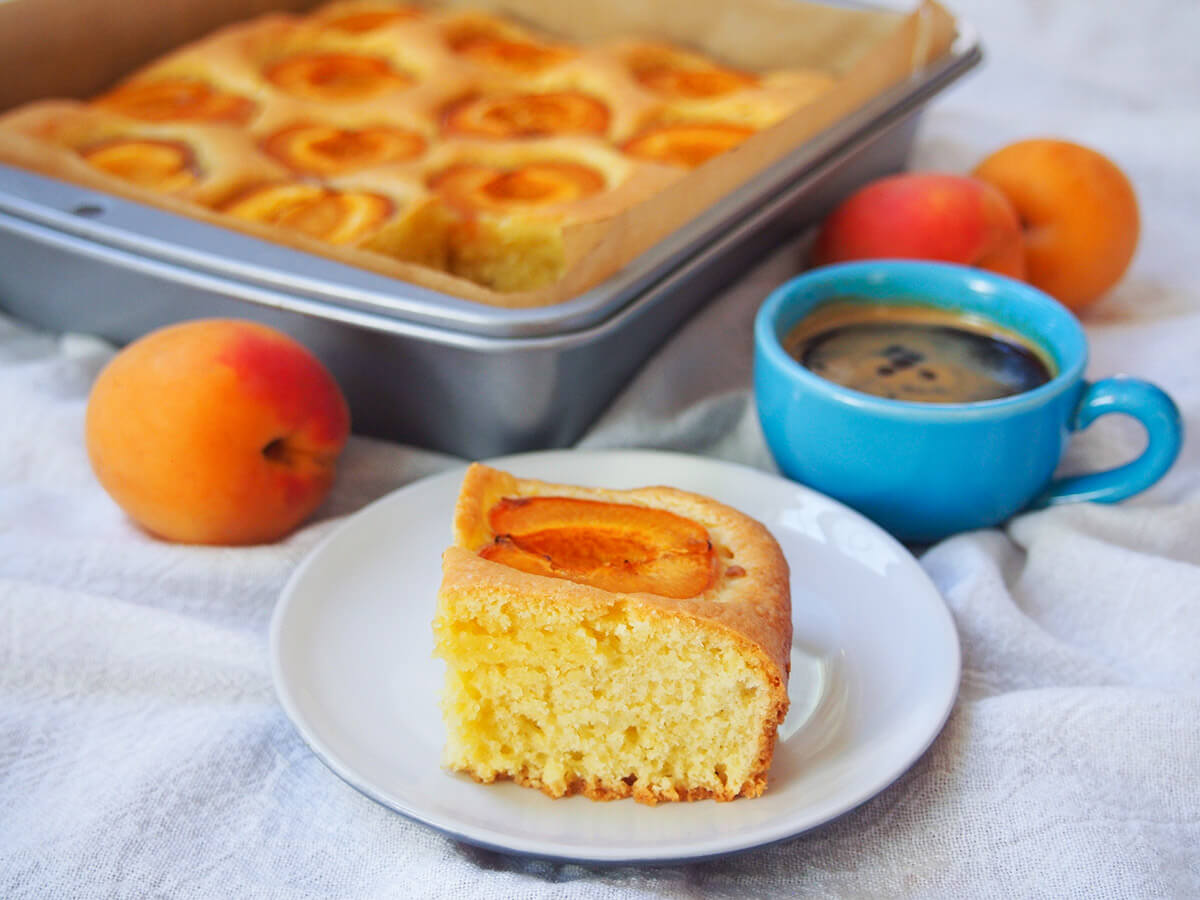 slice of apricot cake marillenkuchen on plate in front of tin with rest of cake, coffee cup to side of tin