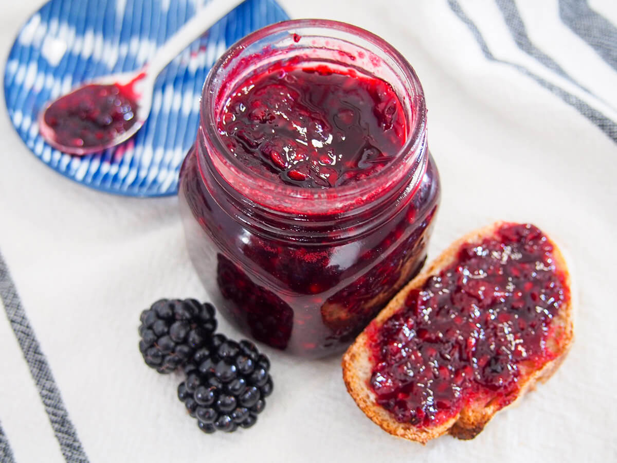 jar of blackberry jam with some on bread to one side and spoonful on small plate behind