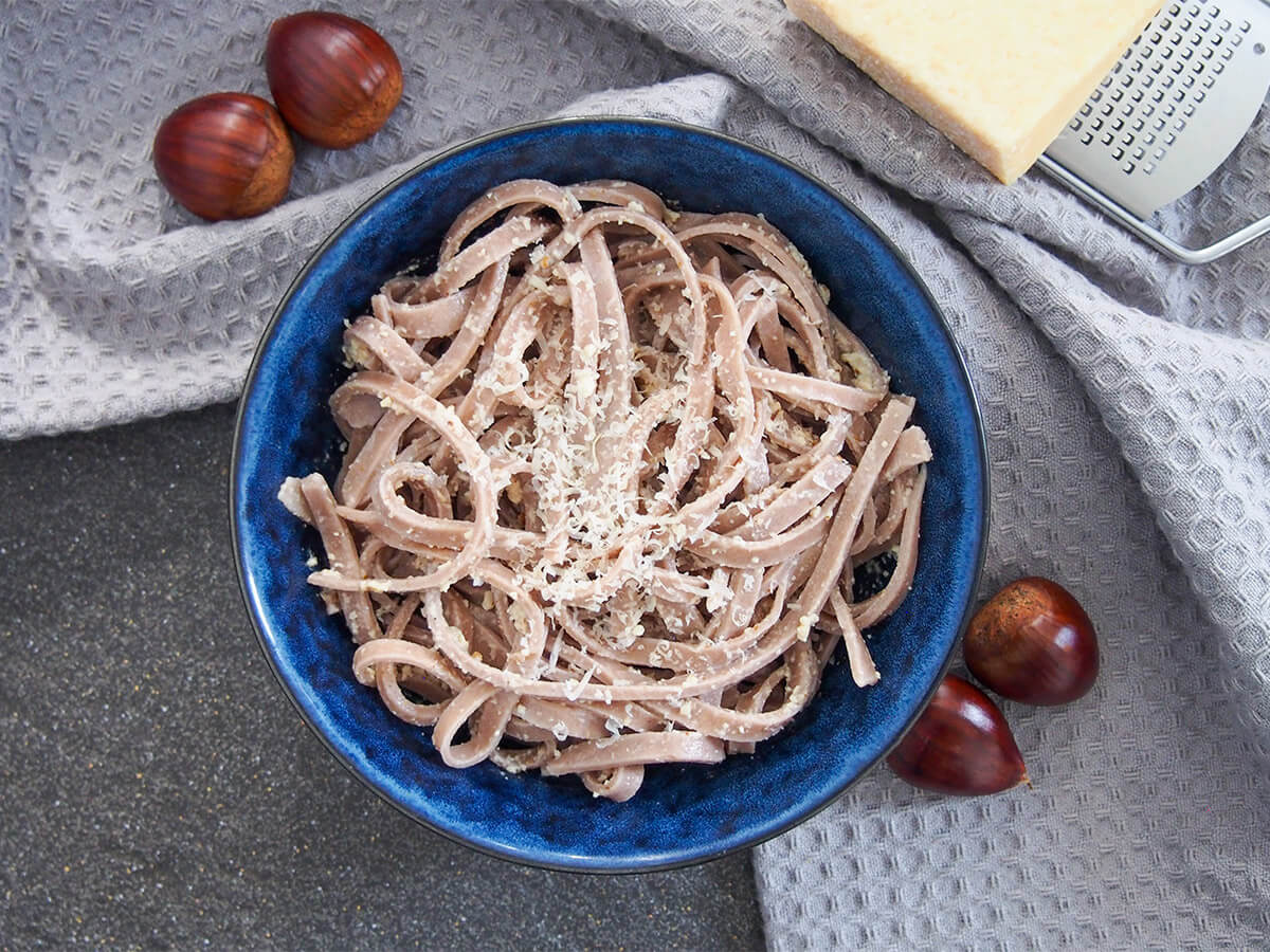 bowl of chestnut pasta with whole chestnuts to side of bowl and cheese on grater above.