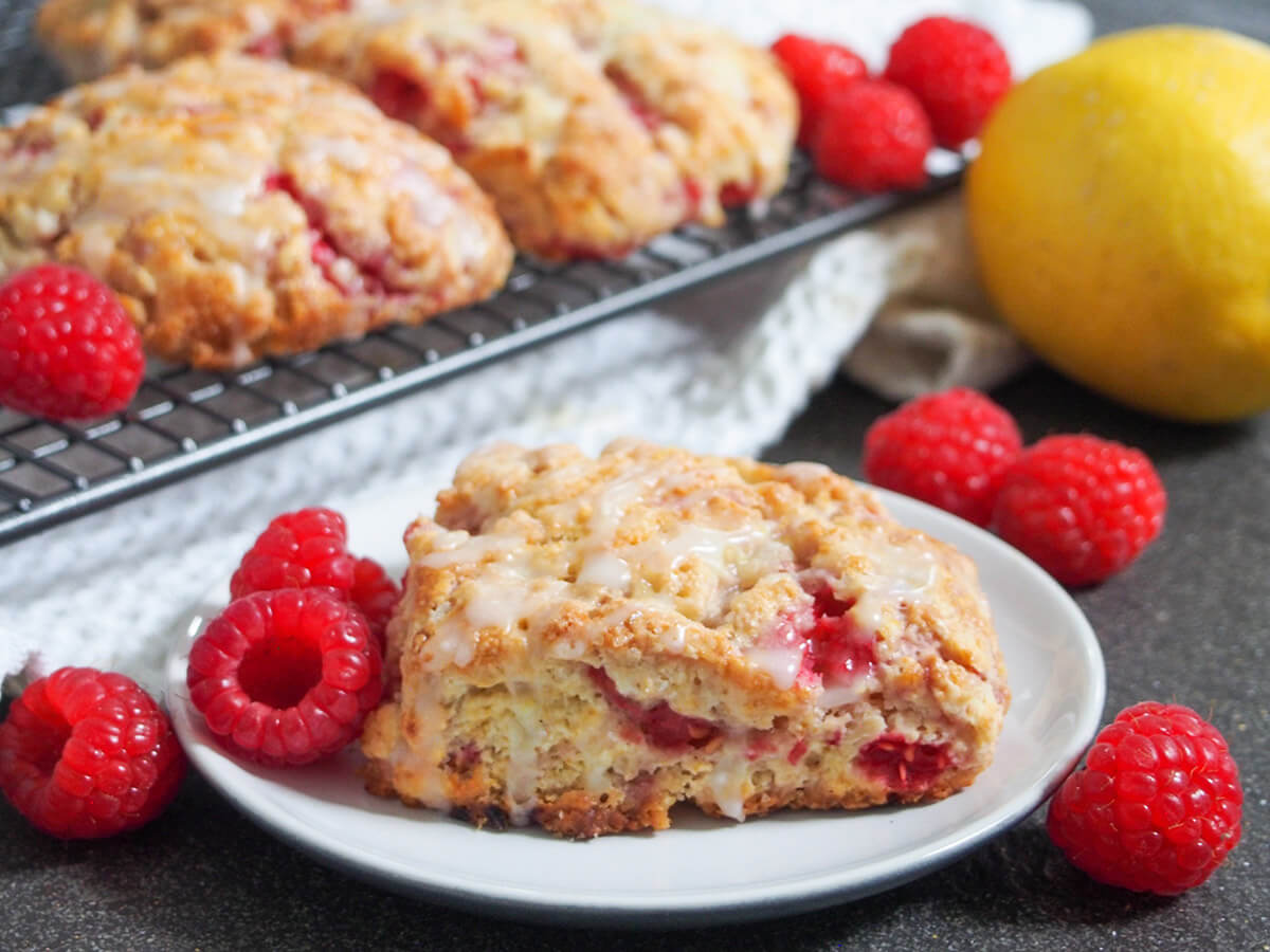 raspberry scone on plate with raspberries around, lemon and more scones behind