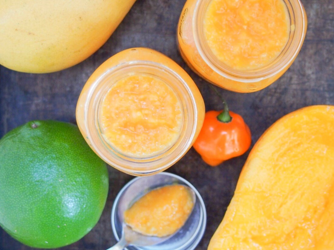 two jars of mango habanero sauce from above with spoon of sauce resting on lid below and lime, chili and mango to side