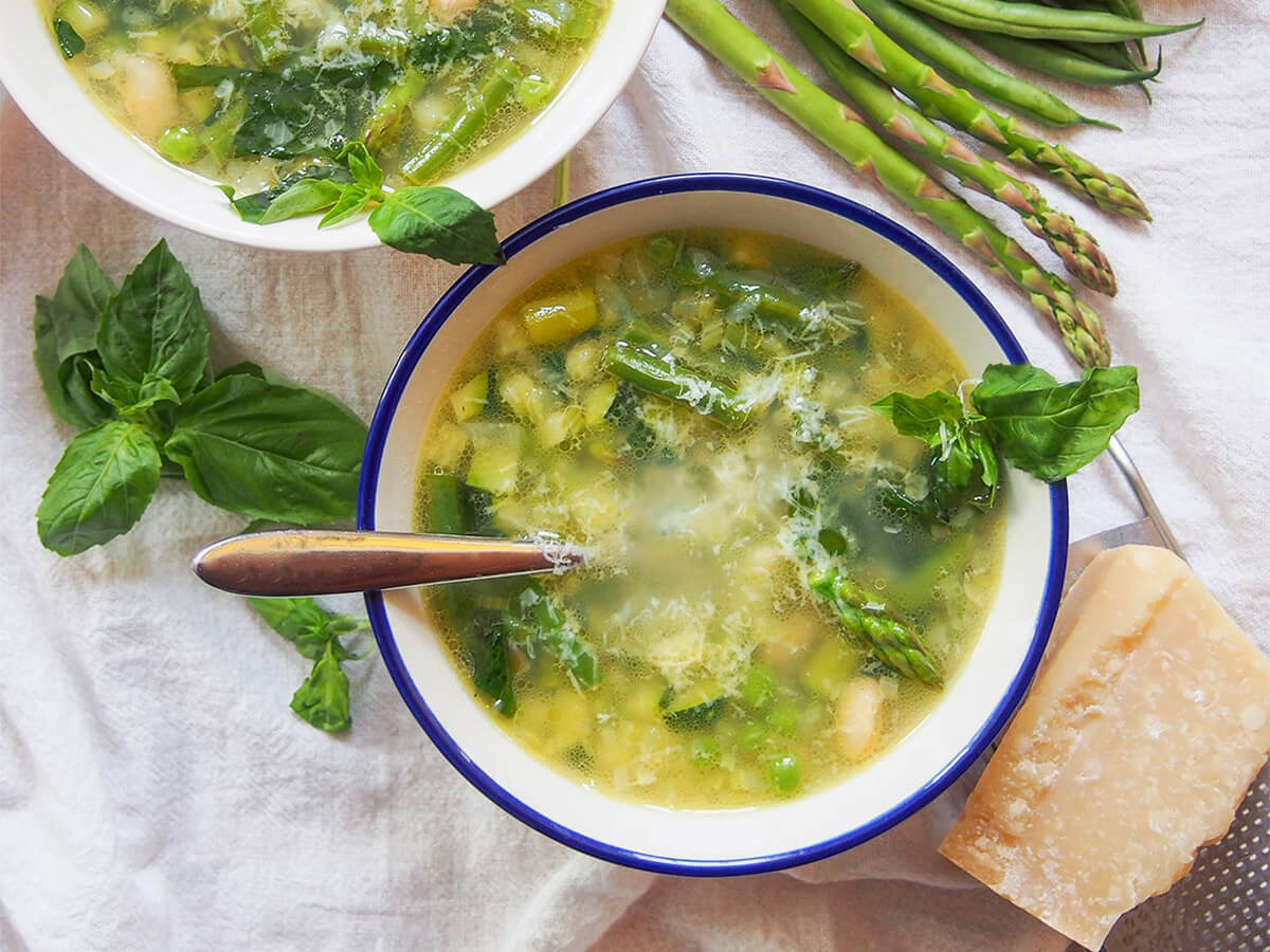 bowl of minestrone primavera with basil to side of bowl and parmesan on other