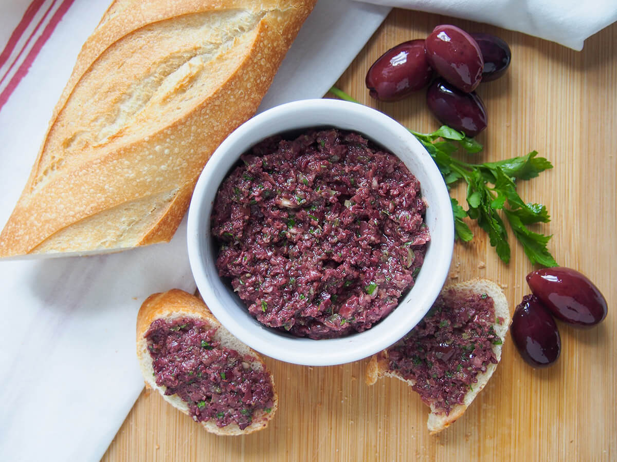 dish of tapenade with part of baguette to side and olives and parsley to other