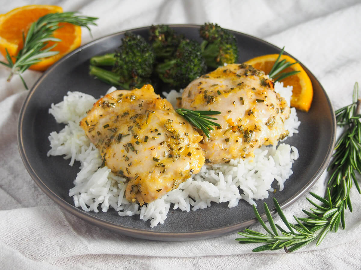 plate with two pieces of orange rosemary chicken over rice with broccoli behind and sprig of rosemary in front of plate