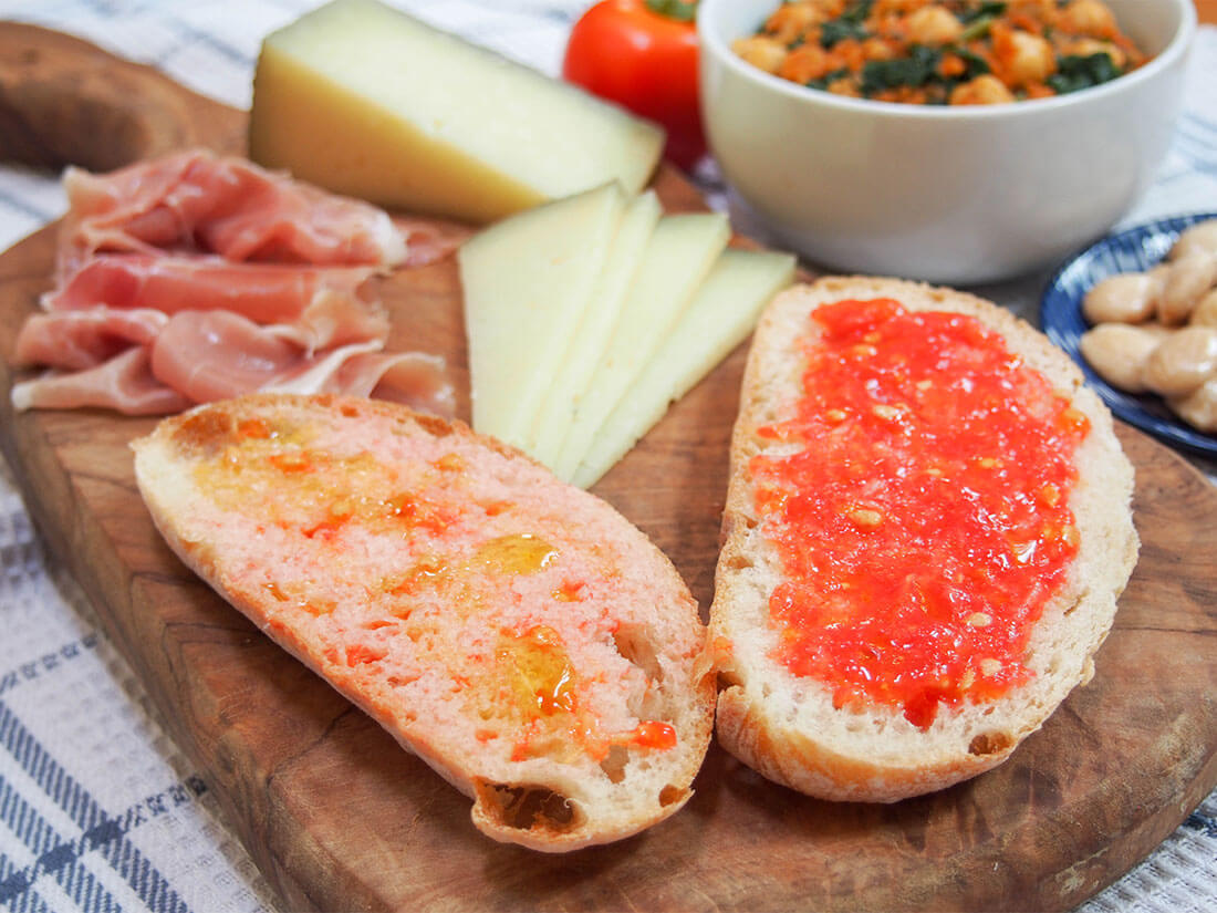 Pan con tomate (pa amb tomaquet) on cheese board with slices of ham and cheese behind