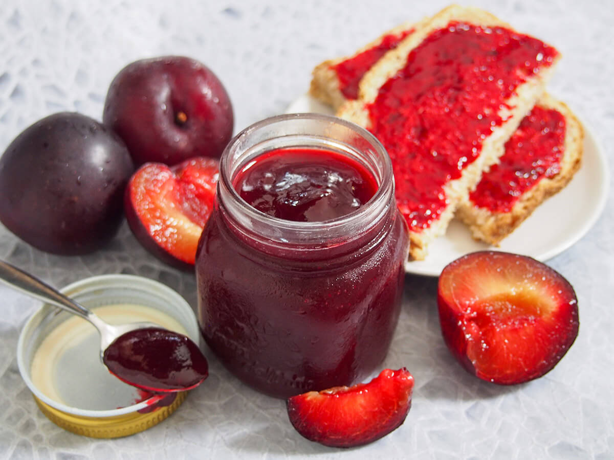 jar of plum jam with plums to side and bread with jam behind, spoon with jam to side of jar