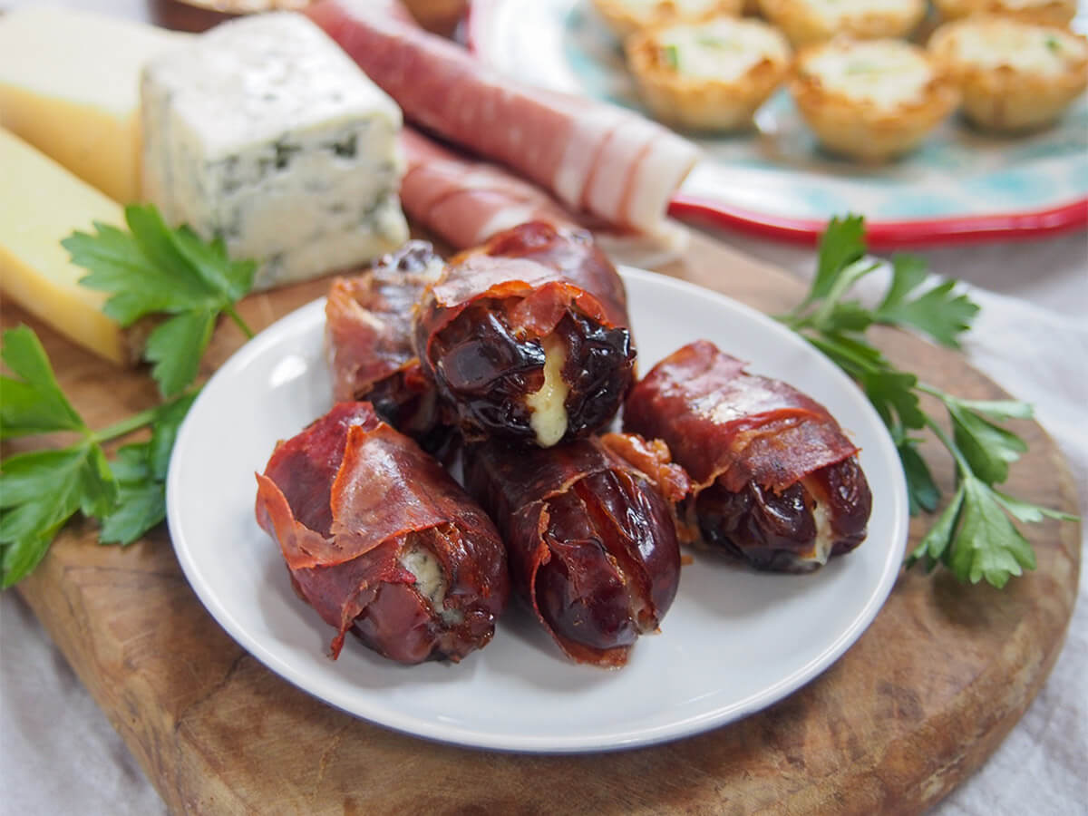 plate of prosciutto wrapped dates on wooden board with cheese and ham behind