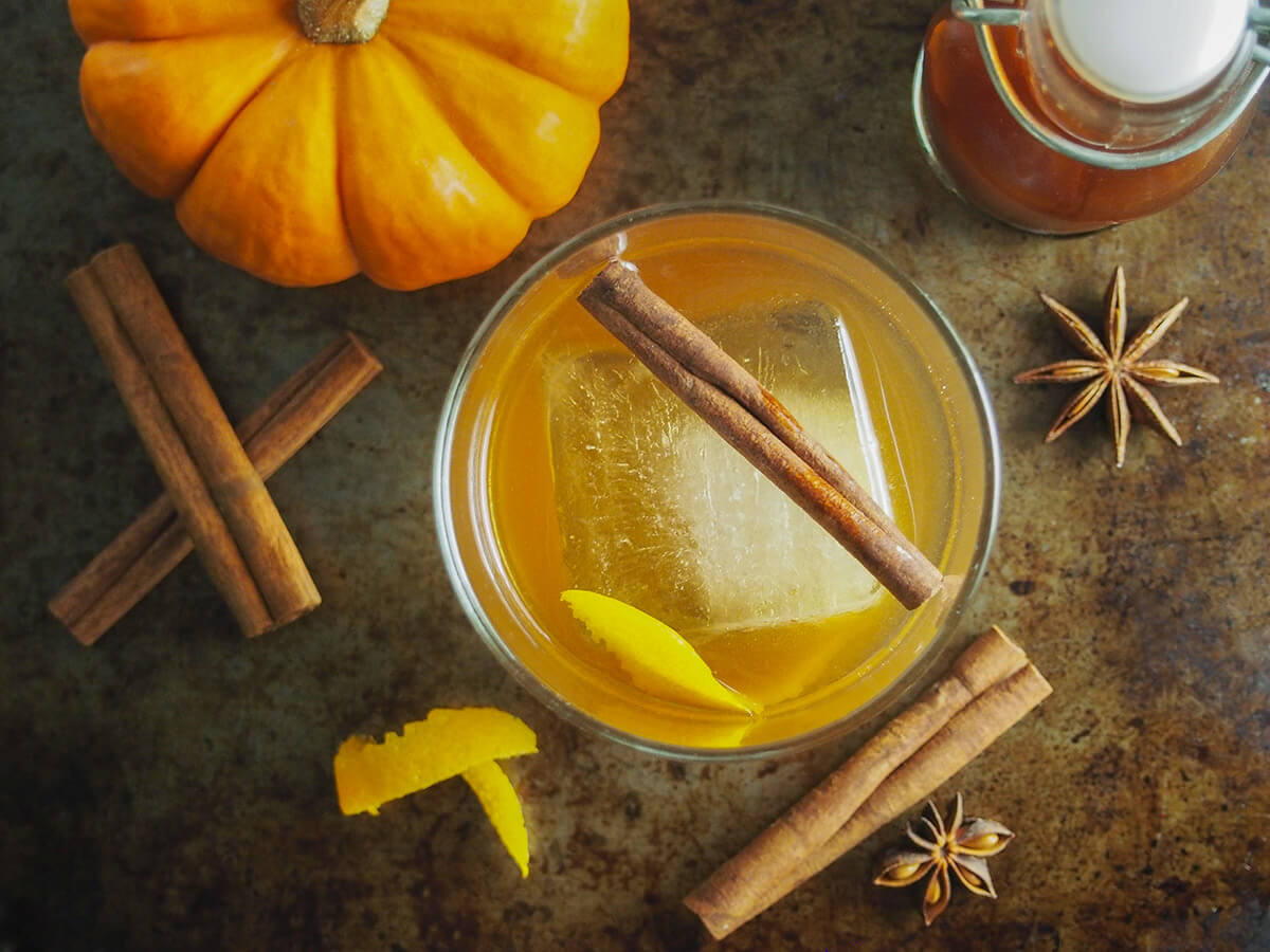 glass of pumpkin spice old fashioned with whole spices, mini pumpkin and bottle of spiced syrup around