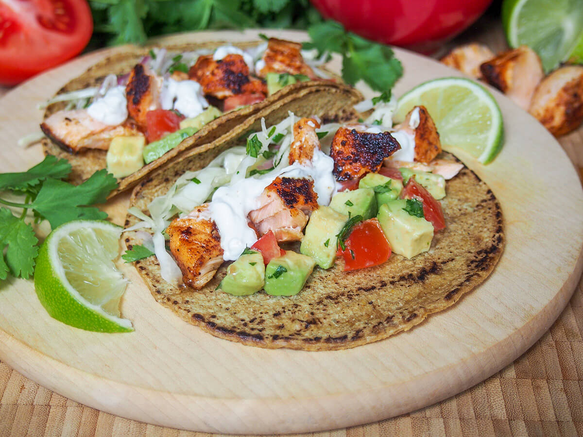 salmon taco with another sitting behind it on a wooden board with slices of lime to either side