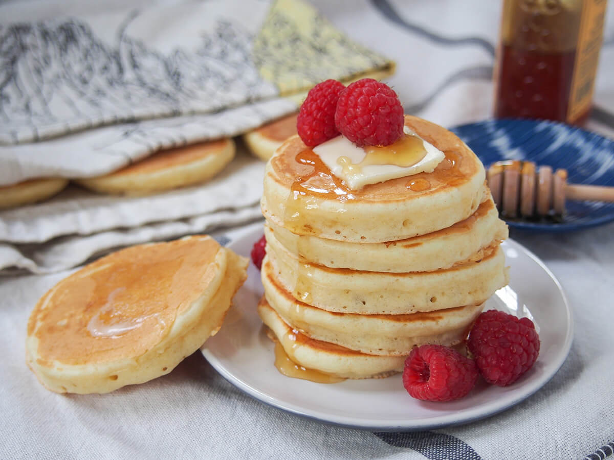 stack of Scotch pancakes on plate with raspberries to side and another pancake resting on side of plate with butter melted on top.