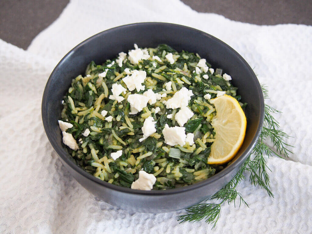 bowl of spanakorizo topped with pieces of feta and lemon slice to side of bowl