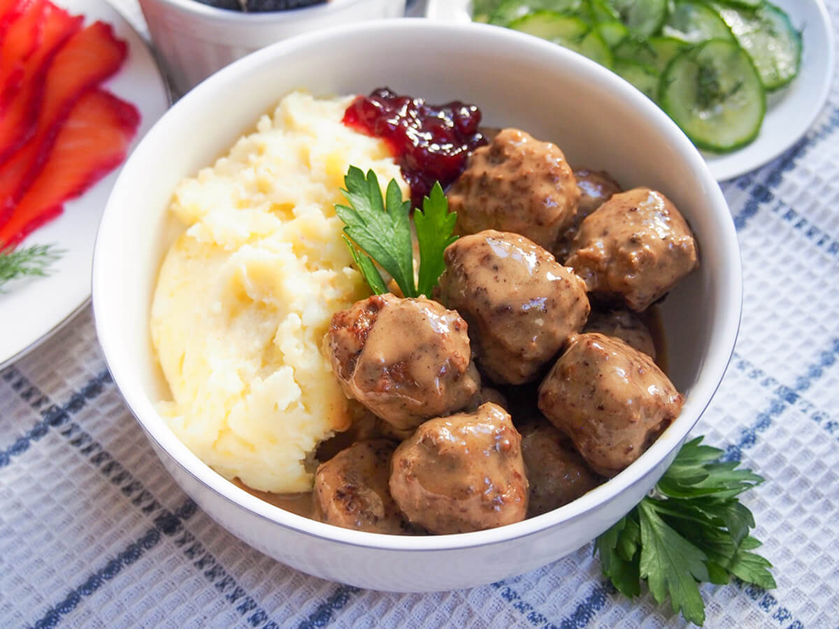 bowl of Swedish meatballs with mashed potato with plates of cucumber and gravlax in background