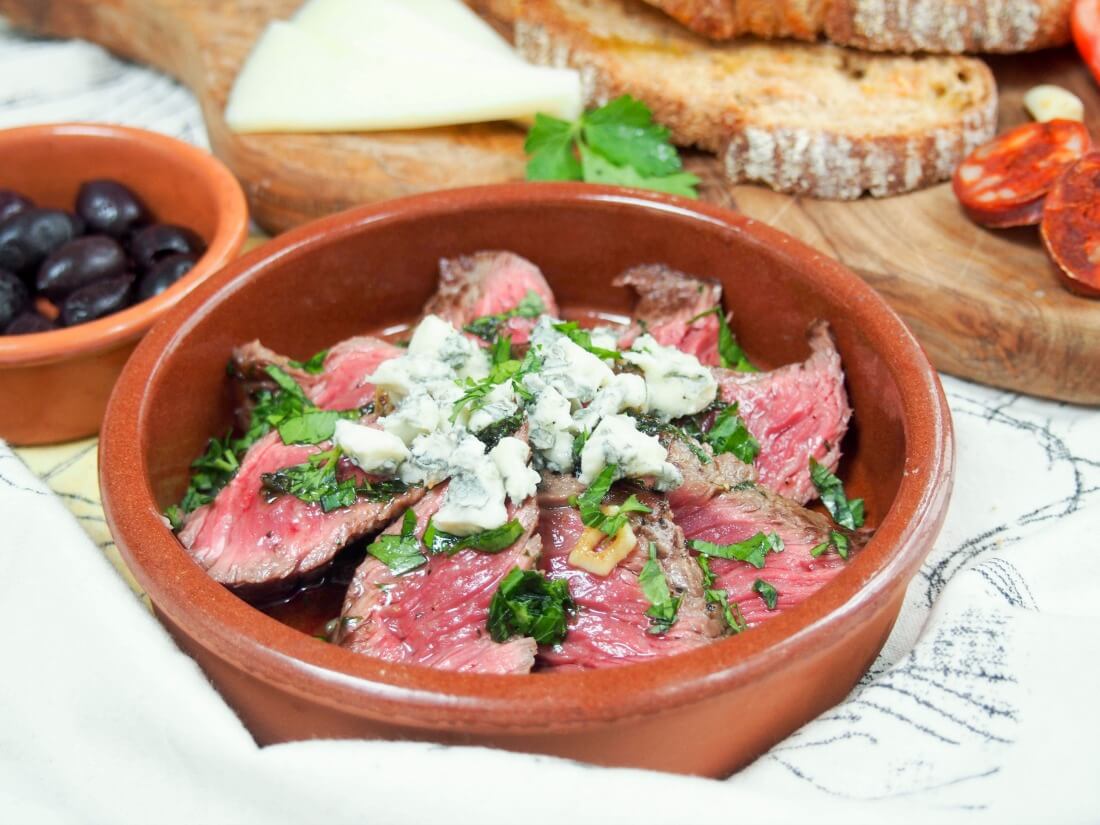 dish with white wine marinated steak topped with blue cheese with some olives and bread behind